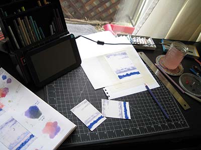 A desk with drawing paper and cards, the paper is still white except for some clouds and trees drawn in pencil crayon. Paints, a sketchbook, and a box of pencil crayons surround the drawings.