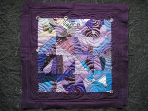 A patchwork sampler with curved quilted lines, embellished with beads and buttons; the edges are still rough.