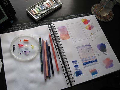 A desk with an open sketchbook, pencil crayons, a box of watercolor tubes. On the right-hand page are color swatches and test drawings.