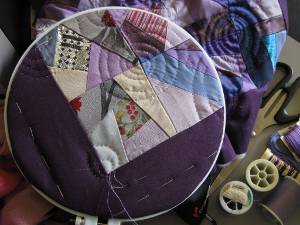 Patchwork sampler stretched on an embroidery hoop, a line of stitching in progress.