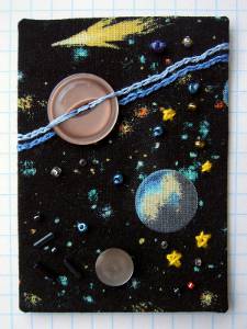 An ATC made with fabric, black background with printed stars and planets, with additional buttons/beads/threads sewn over top.