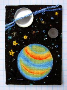 An ATC made with fabric, black background with printed stars and planets, with additional buttons/beads/threads sewn over top.